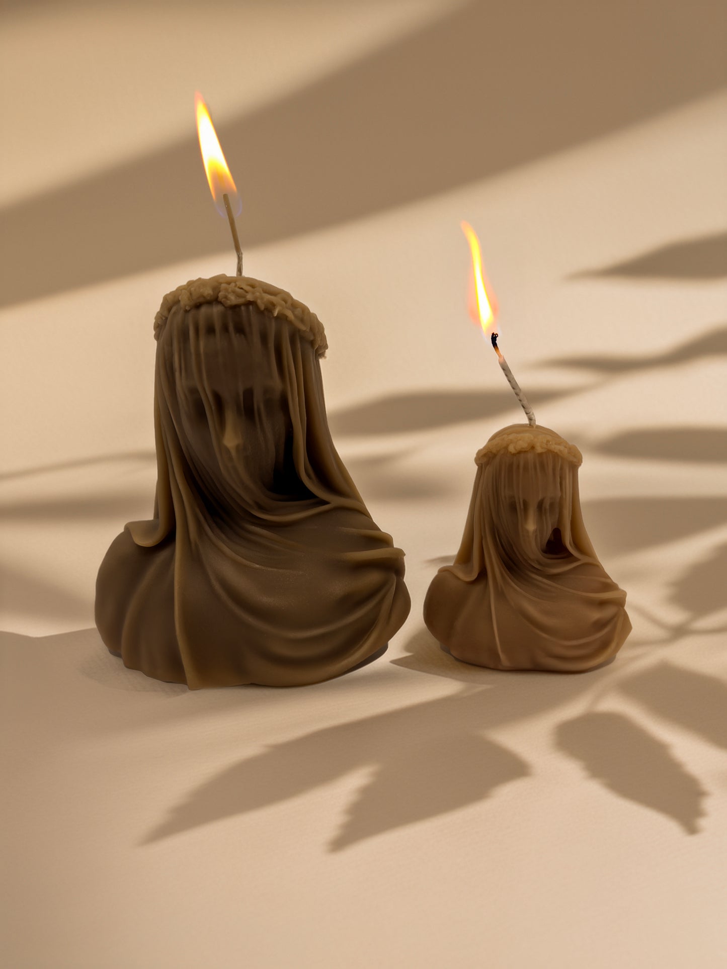 Two brown wax figures with hooded veils and lit candles on a beige background