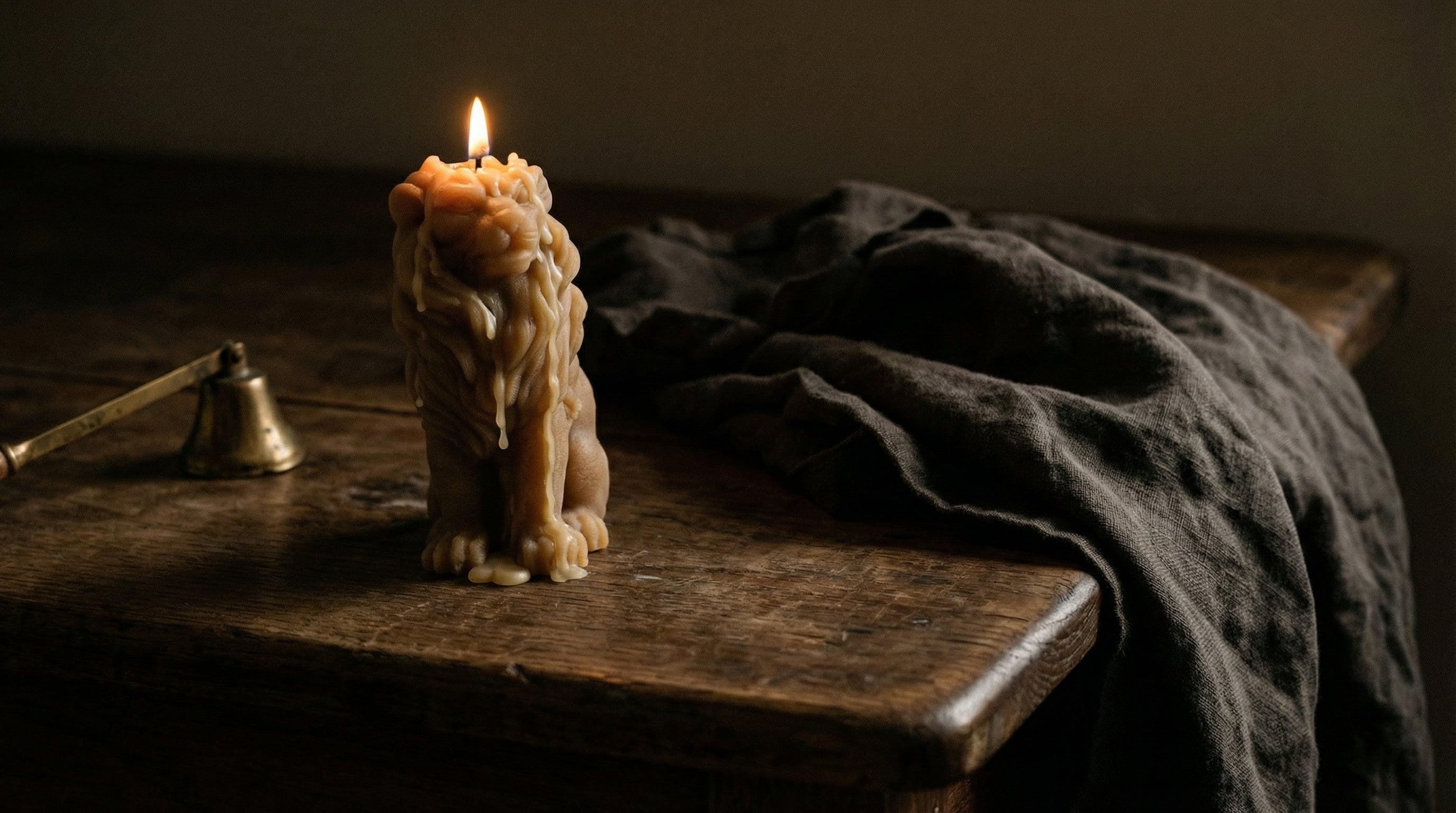 Candle in the shape of a lion on a wooden surface with a dark cloth and bell in the background.