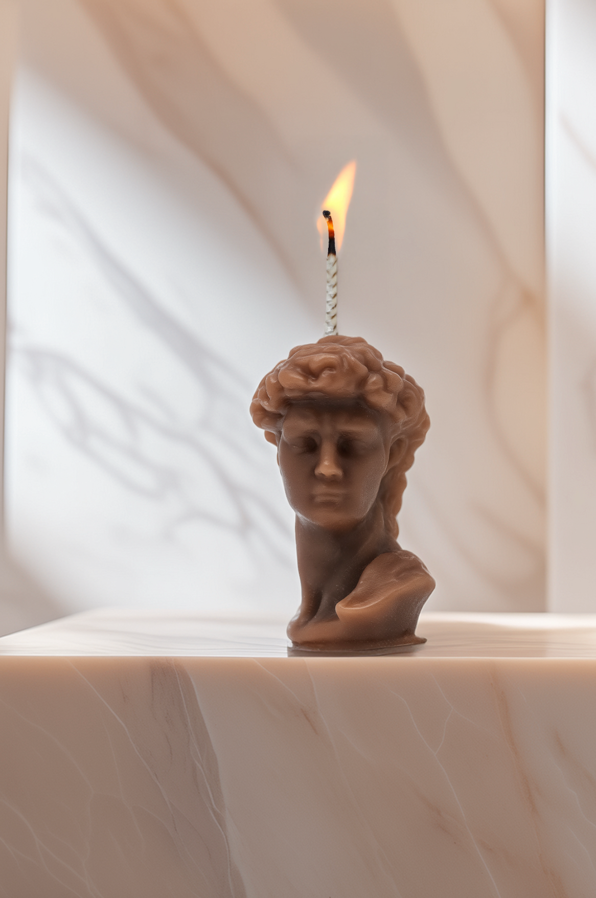 Candle shaped like a classical sculpture bust on a marble surface with a white curtain background