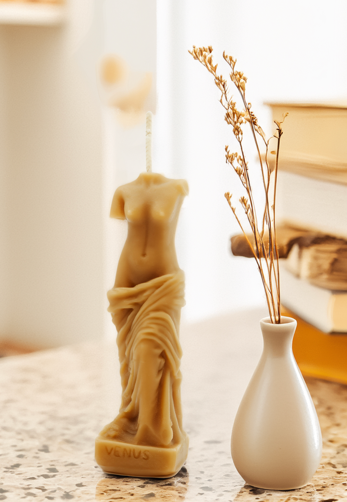 Candle shaped like a classical statue next to a white vase with dried plants on a textured surface.